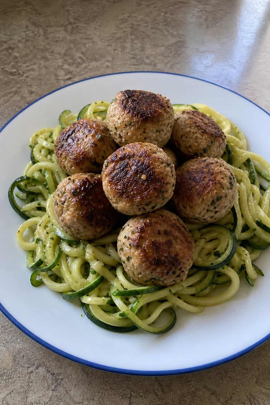 A plate of turkey-chicken meatballs served with zucchini noodles on a wooden table with fresh ingredients in the background.