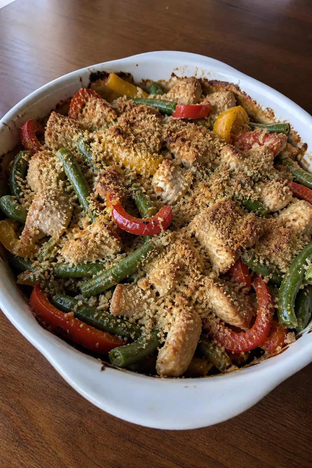A baked turkey and green bean casserole with colorful red and orange bell pepper strips visible through the crispy topping, in a white ceramic dish on a wooden table.