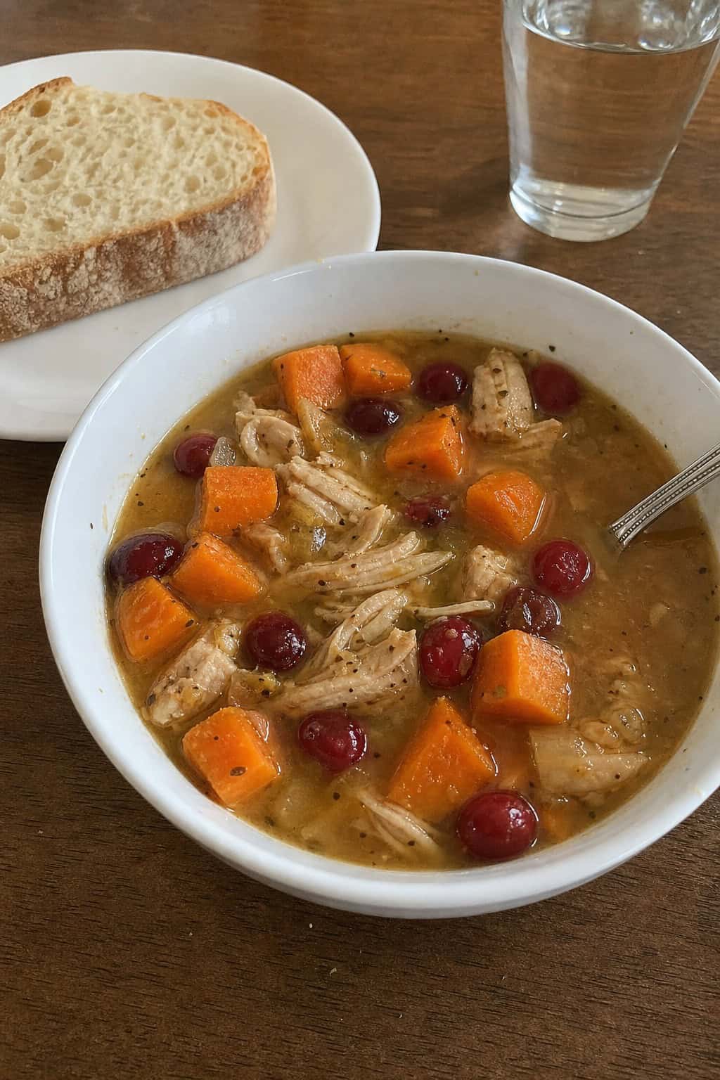 A bowl of turkey, sweet potato, and cranberry soup on a wooden table with fresh ingredients and bread nearby.