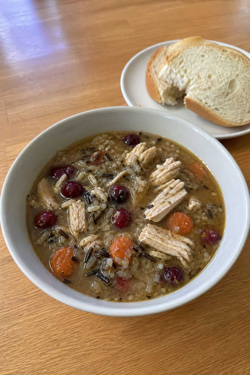 A bowl of turkey, wild rice, and cranberry soup garnished with herbs on a wooden table with ingredients around it.