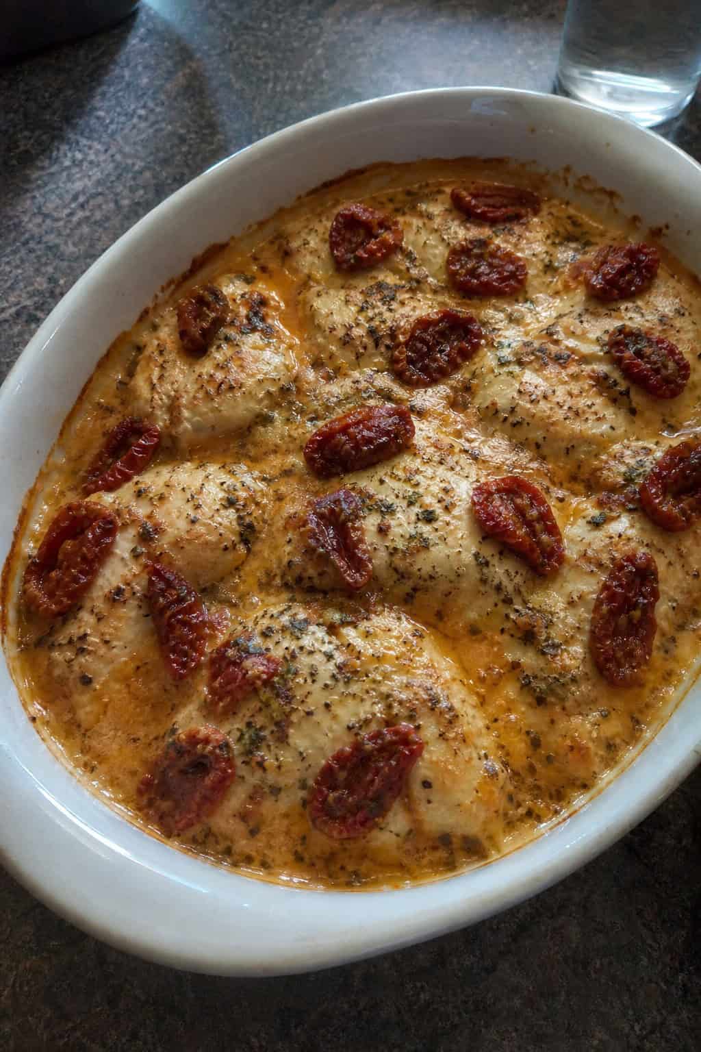 A baked chicken and sun-dried tomato casserole with pasta in a cream sauce, topped with melted mozzarella and Parmesan in a ceramic dish on a wooden table.