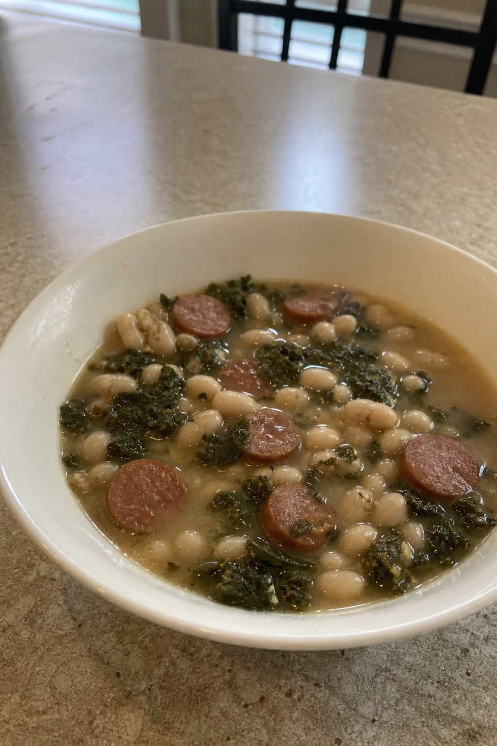 A crockpot filled with steaming Tuscan white bean, kale, and sausage stew on a wooden countertop surrounded by fresh ingredients.