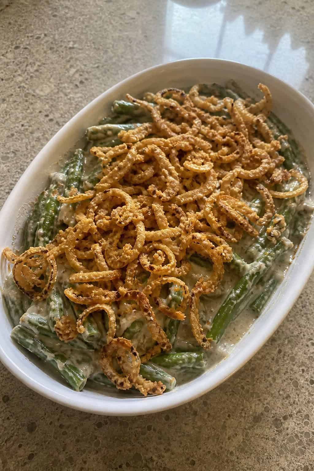 A vegan green bean casserole topped with golden crispy fried onions in a white ceramic dish on a wooden table, with fresh green beans and a small pile of raw cashews arranged beside it.