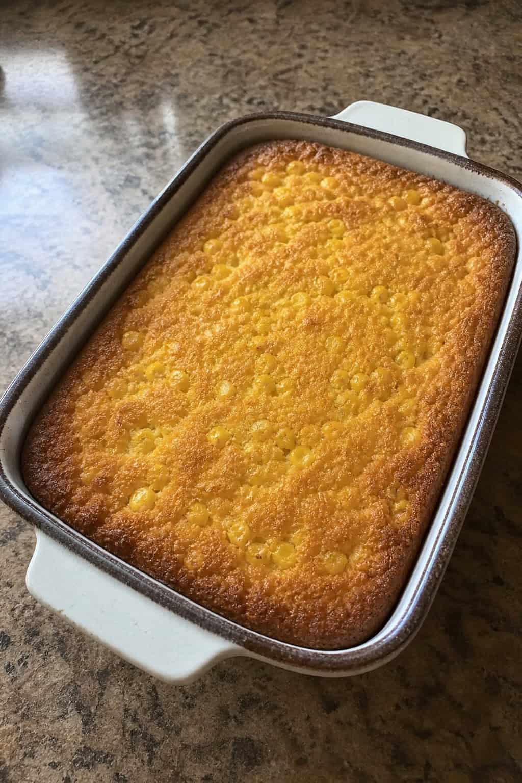 A golden baked vegan corn casserole in a white ceramic dish on a wooden table with fresh corn ears and small bunches of herbs arranged around the dish.