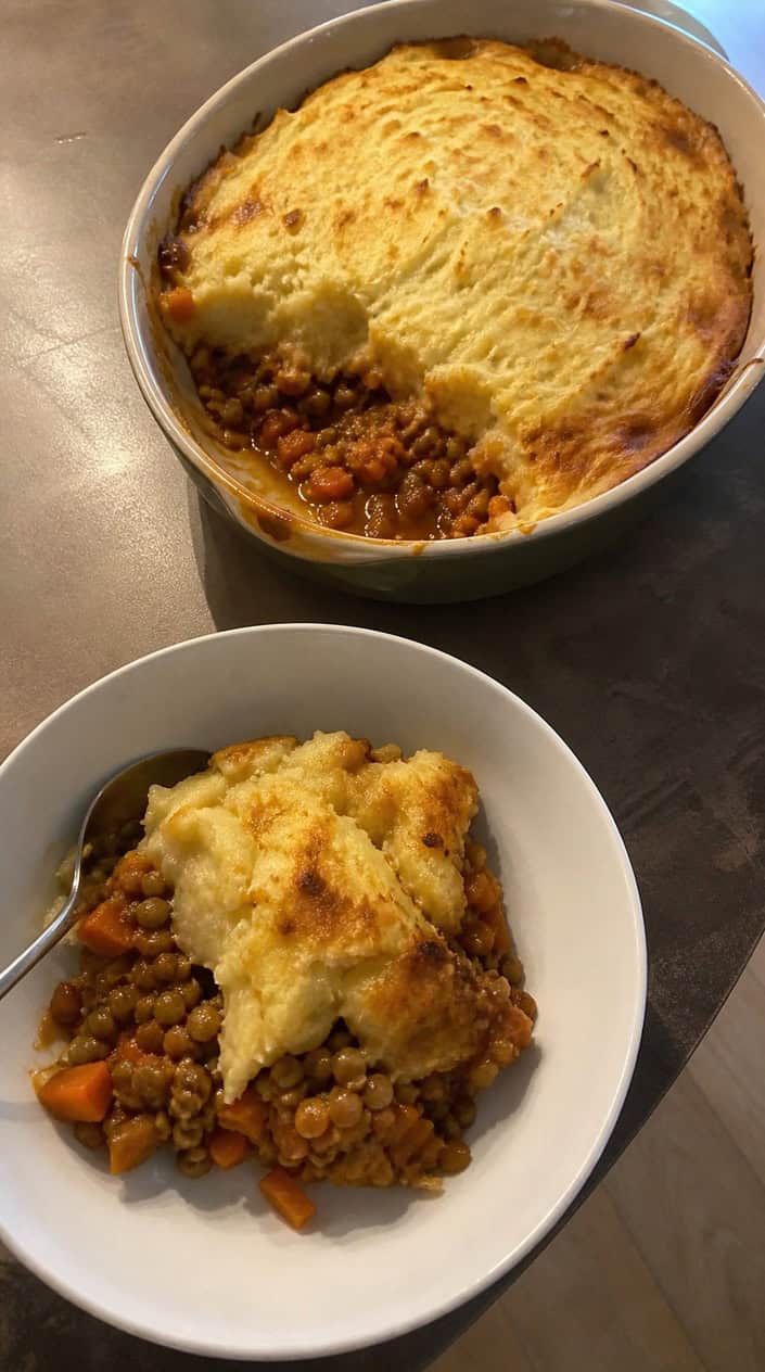 A freshly baked vegetarian lentil shepherd’s pie with golden mashed potato topping on a wooden table, accompanied by a green salad and a glass of water.