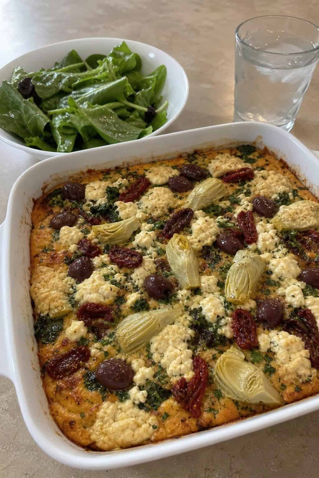 A freshly baked vegetarian Mediterranean hashbrown casserole in a ceramic dish on a wooden table, garnished with fresh herbs, next to a bowl of mixed greens and a glass of water with lemon.