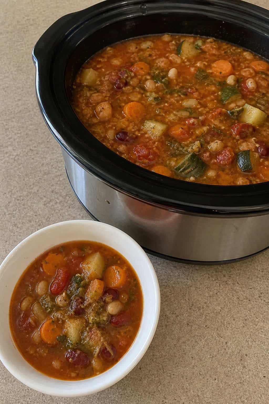 A crockpot filled with colorful vegetarian minestrone soup next to a bowl of green pesto and fresh vegetables on a kitchen counter.