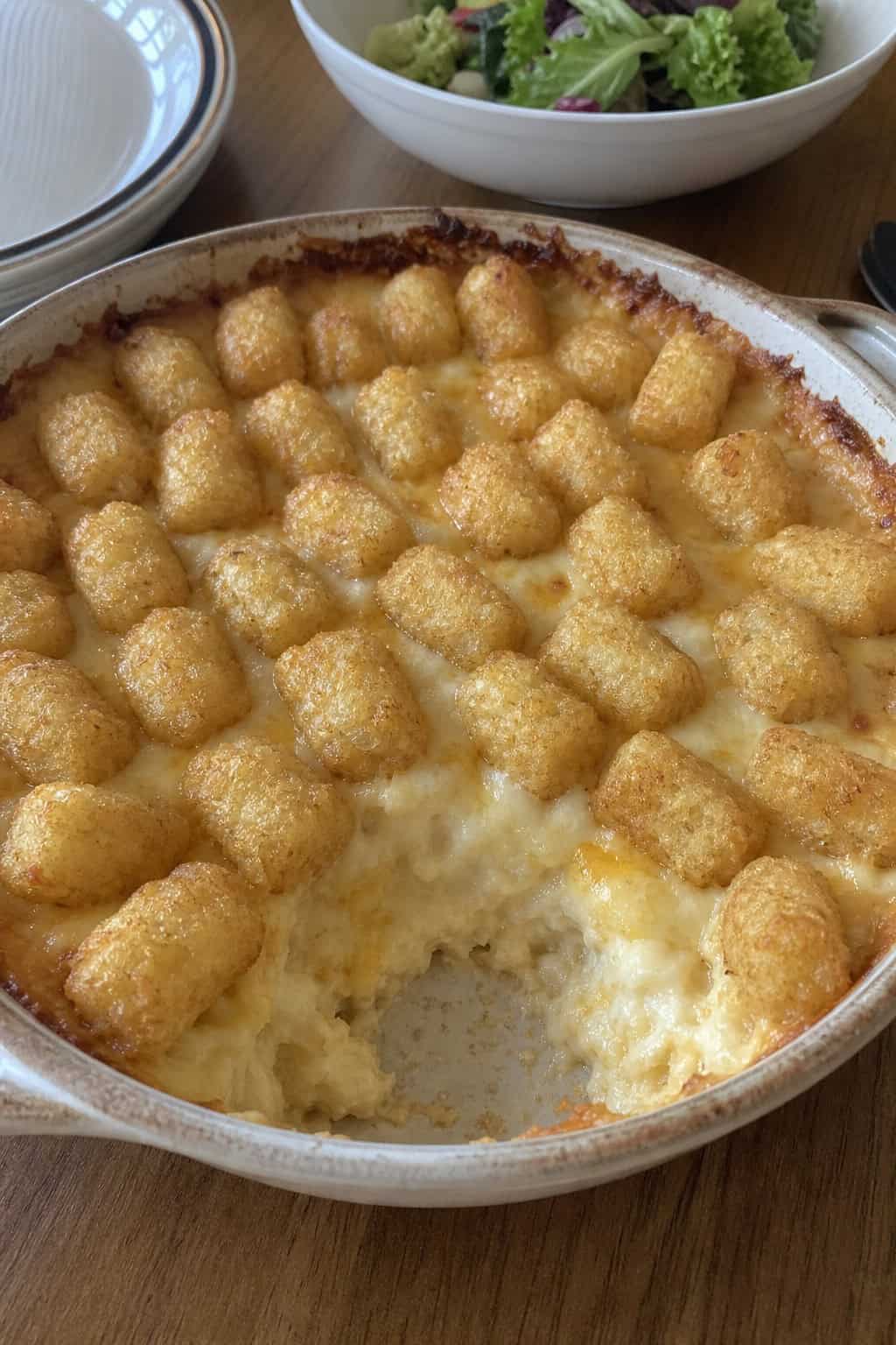 A baked vegetarian seven-cheese tater tot casserole in a cream ceramic dish, with a golden panko-and-tot crust on top and a scattering of fresh chopped parsley.