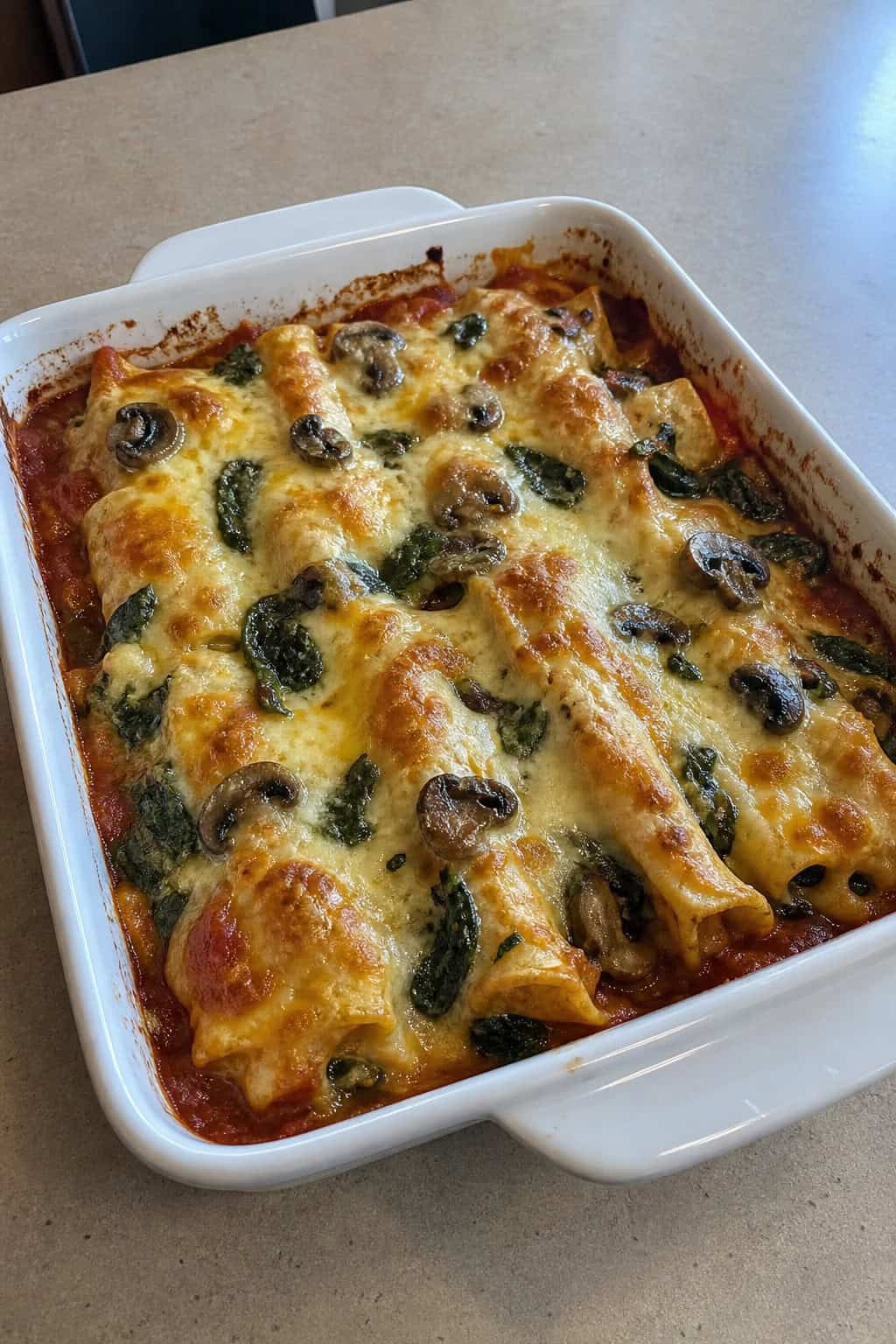A baked spinach and mushroom enchilada casserole in a deep ceramic dish with browned Monterey Jack cheese on top, with fresh spinach leaves and sliced cremini mushrooms on the wooden table beside it.