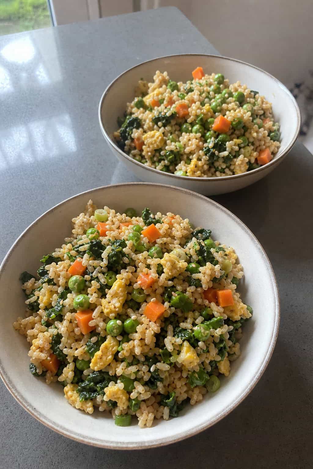 Two bowls of colorful veggie fried rice with greens on a wooden table, accompanied by chopsticks and a small bowl of soy sauce.