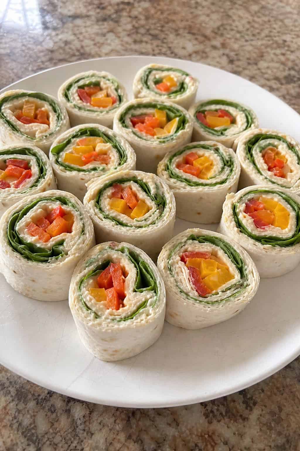 Plate of colorful veggie hummus pinwheels showing bright red and yellow bell pepper strips, shredded orange carrot, and green spinach inside sliced flour tortilla rolls on a wooden board.