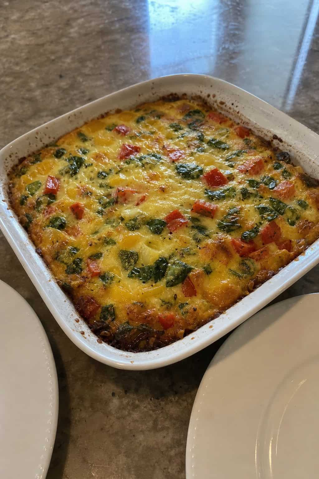 A baked vegetable hashbrown casserole with visible red and green bell pepper pieces and wilted spinach, in a glass baking dish on a wood table surrounded by loose fresh vegetables.