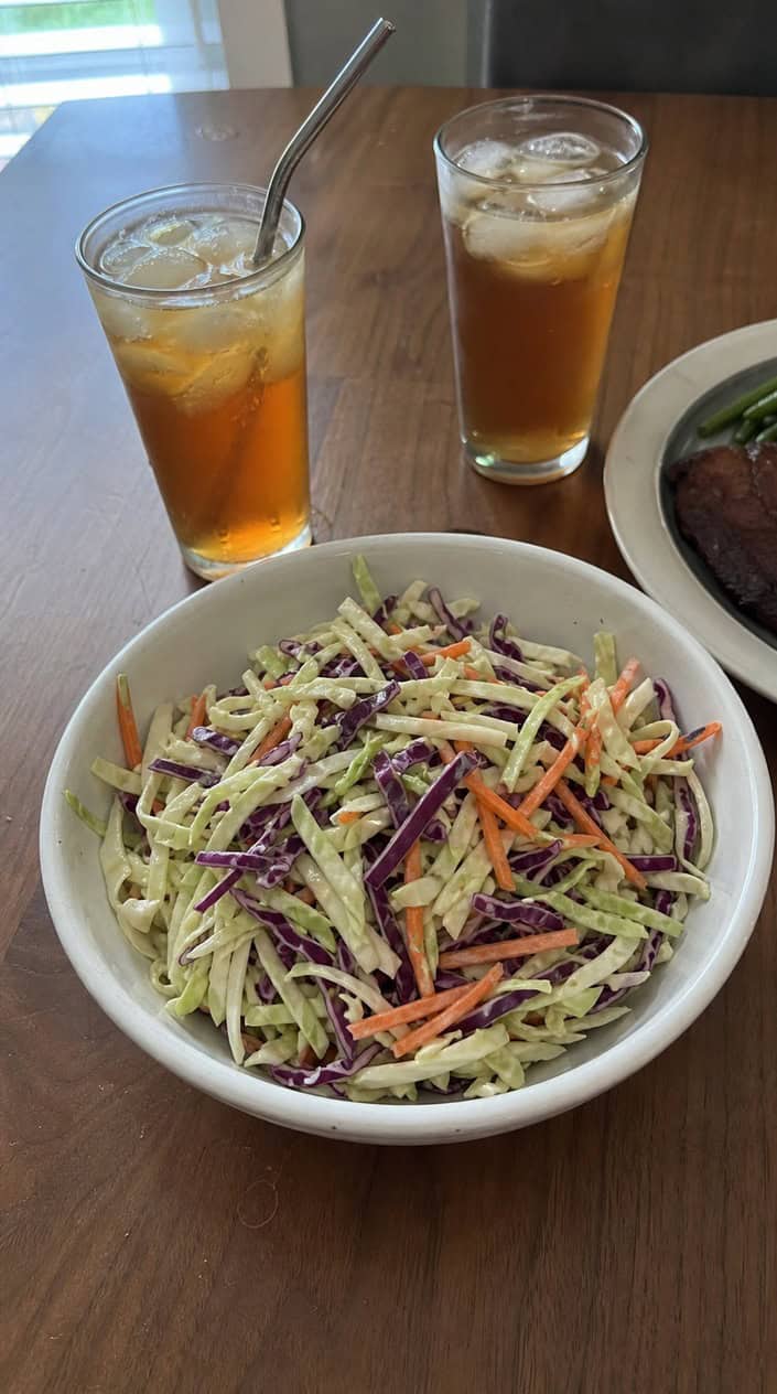 A bowl of vinegar-based Carolina coleslaw on a wooden picnic table with a checkered napkin and a glass of iced tea in an outdoor BBQ setting.