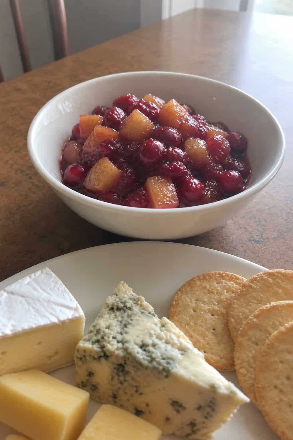 A bowl of warm apple-cranberry compote on a wooden cheese board with various cheeses, nuts, and crackers arranged around it.
