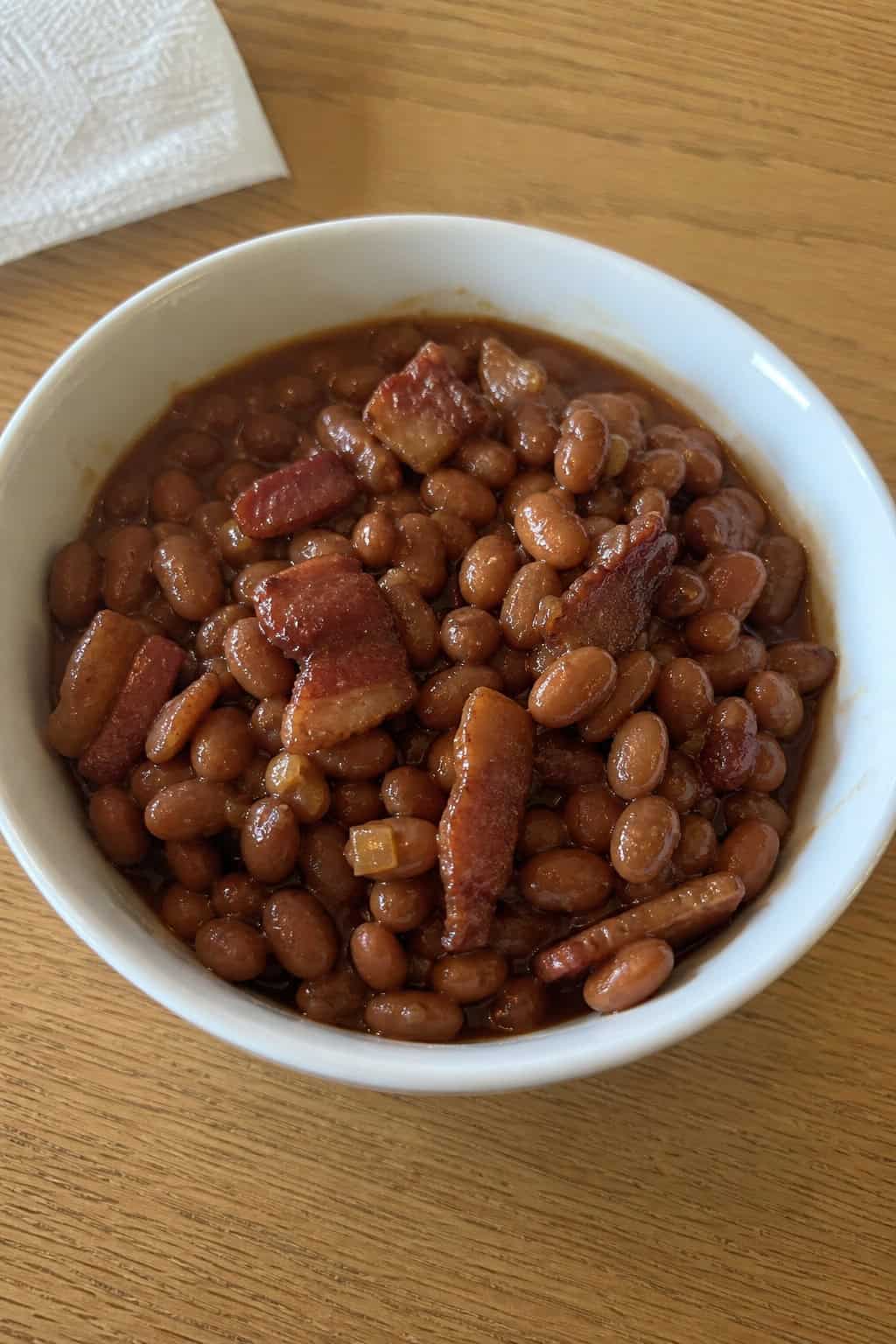 A bowl of warm pinto beans cooked with bacon on a wooden table, surrounded by fresh ingredients.