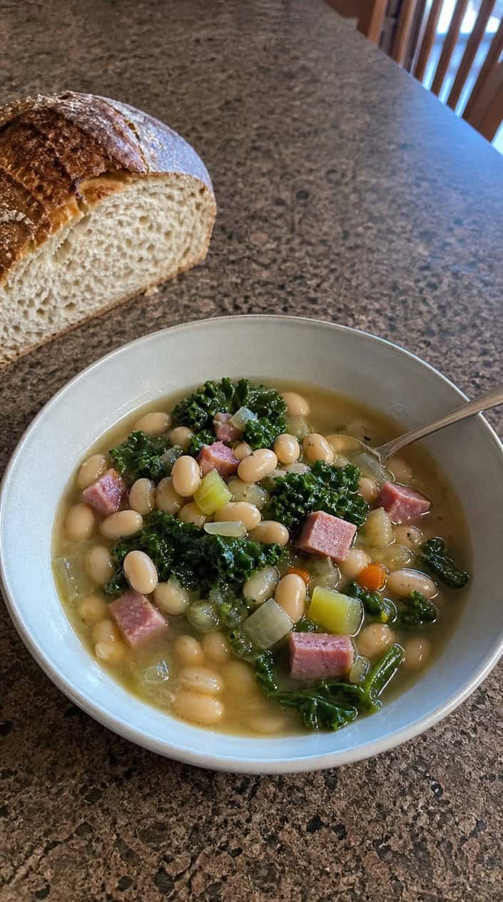 A bowl of white bean and kale ham soup with chunks of ham and leafy kale on a wooden table with a spoon and bread nearby.