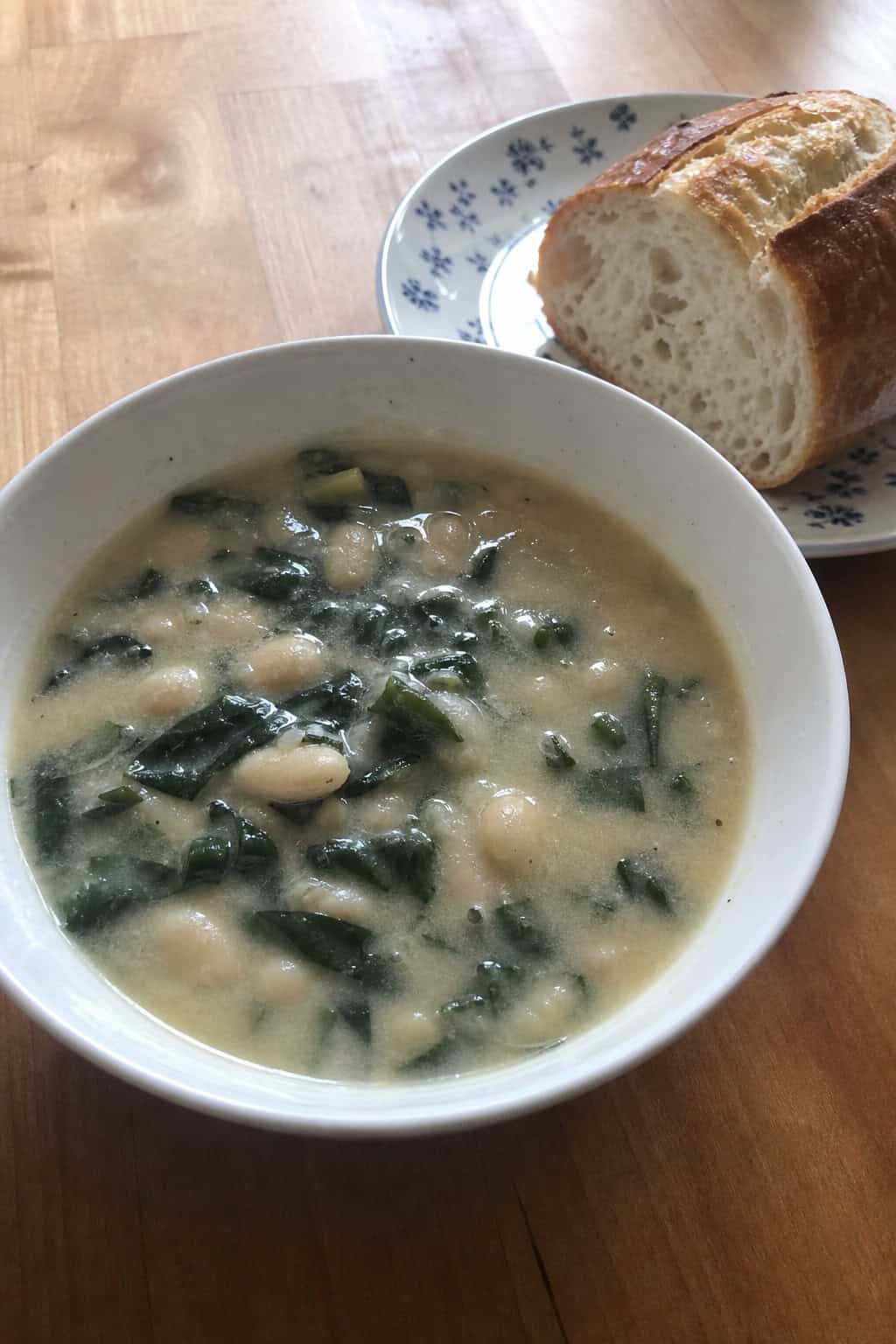 A bowl of white bean, spinach, and rosemary soup on a wooden table with a spoon and bread nearby.