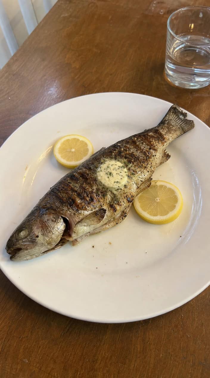 Whole grilled trout garnished with lemon slices, dill, and butter on a wooden platter outdoors.