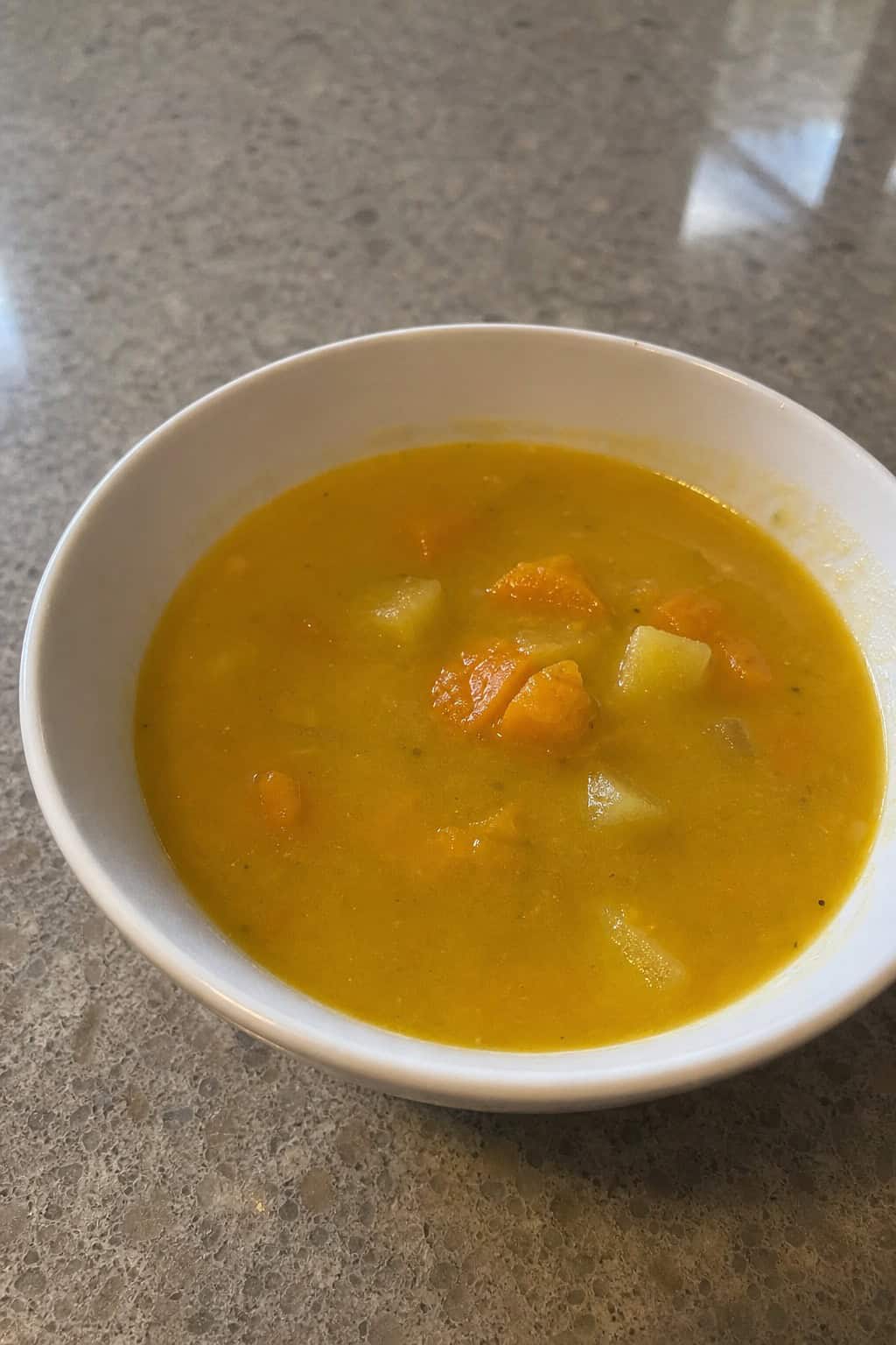 A crockpot filled with steaming winter squash, apple, and ginger soup on a wooden countertop surrounded by fresh squash, apples, and ginger slices.