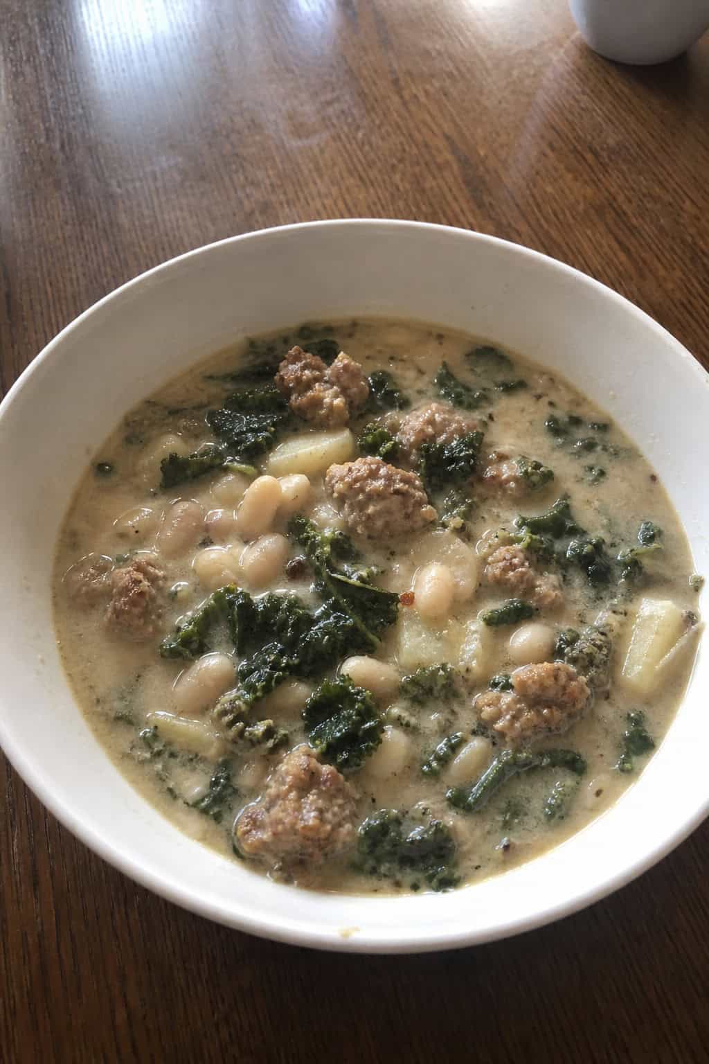 A bowl of Zuppa Toscana soup with kale and white beans on a wooden table surrounded by fresh ingredients and bread.
