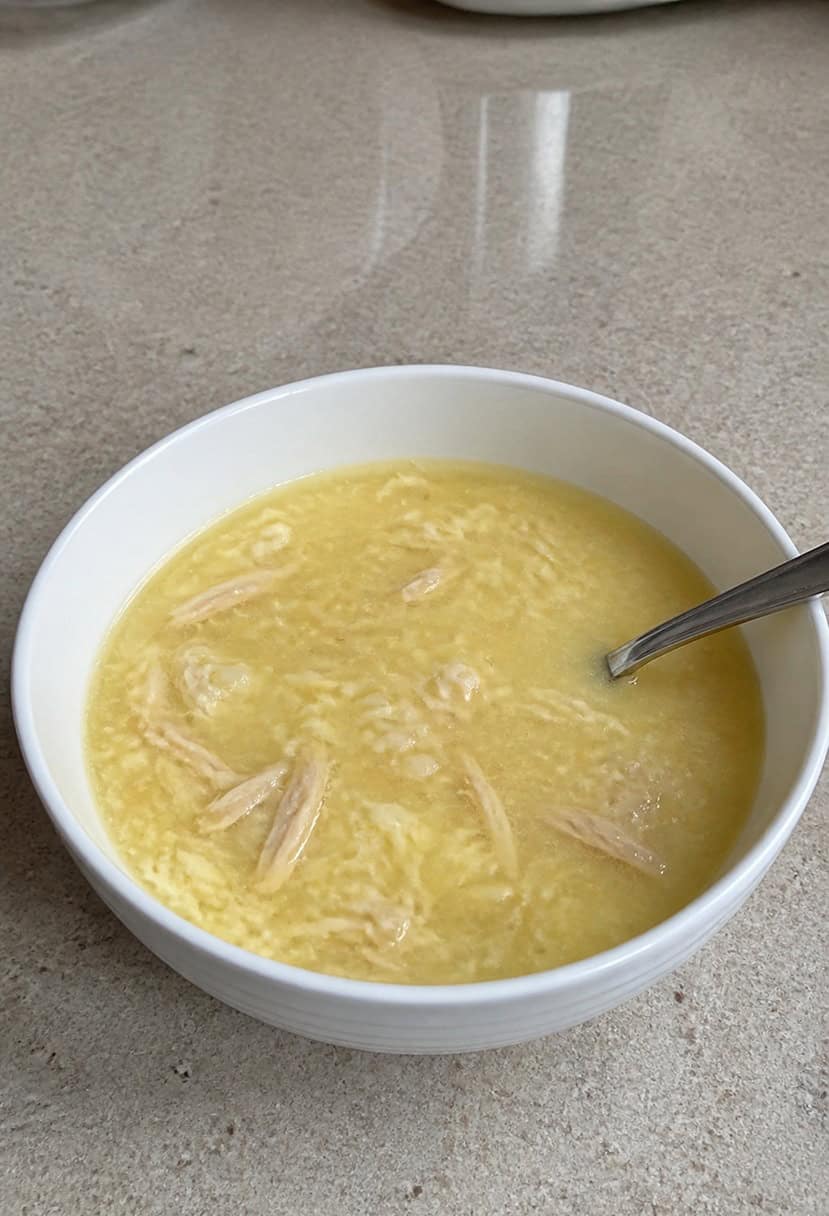A steaming bowl of Avgolemono soup surrounded by fresh ingredients and a spoon on a rustic wooden table