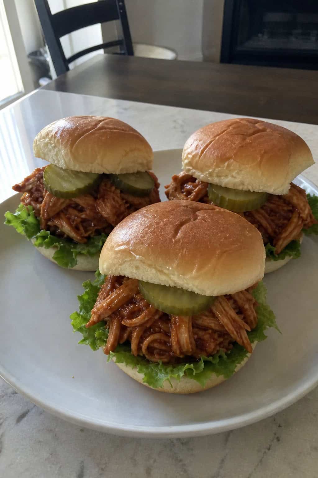 Close-up of BBQ pulled chicken sliders with lettuce and pickles on a wooden board, accompanied by a bowl of BBQ sauce and potato chips.
