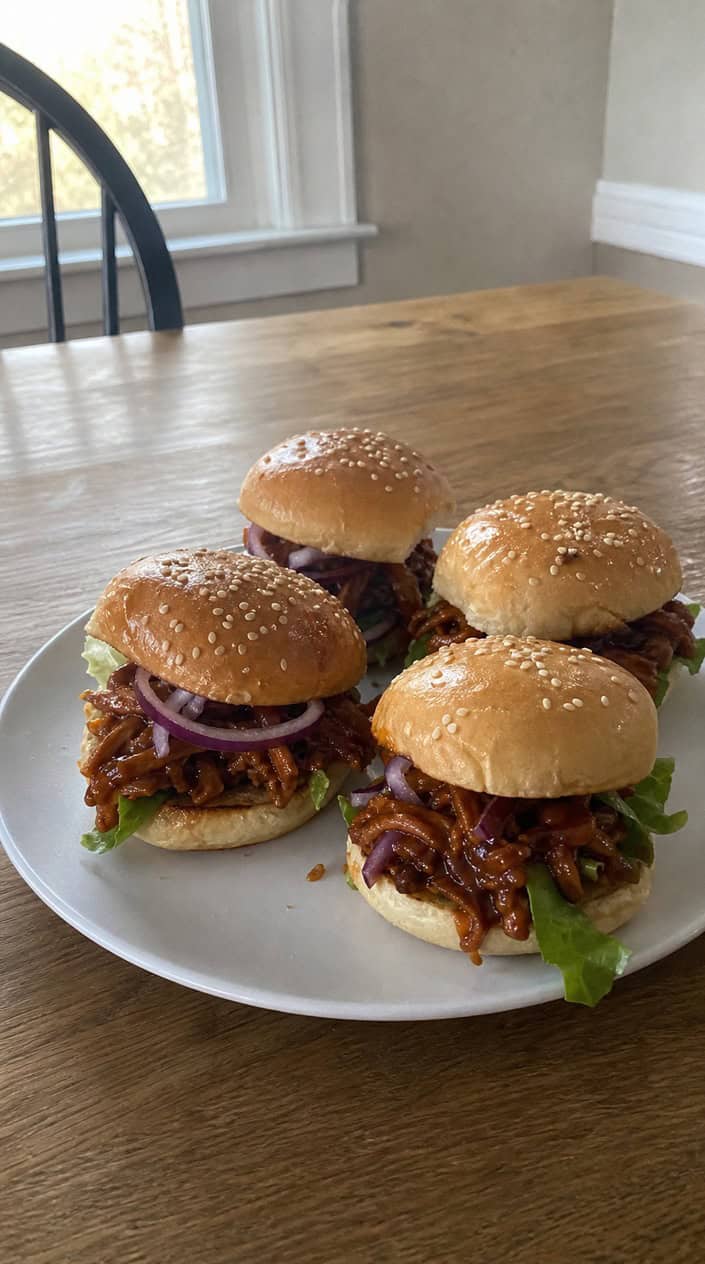 Plate of BBQ pulled jackfruit sliders with lettuce and onions on a wooden table, accompanied by side dishes and a beverage in a casual game day setting.