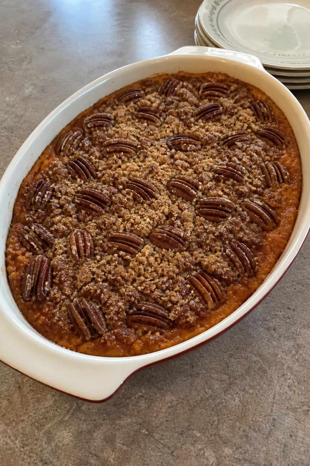 A brown sugar and pecan topped sweet potato casserole in a ceramic dish on a wooden table with autumn decorations around it.