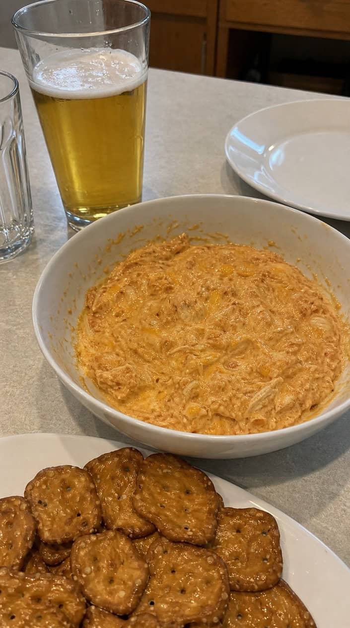 A bowl of buffalo chicken dip surrounded by pretzel chips on a wooden table.