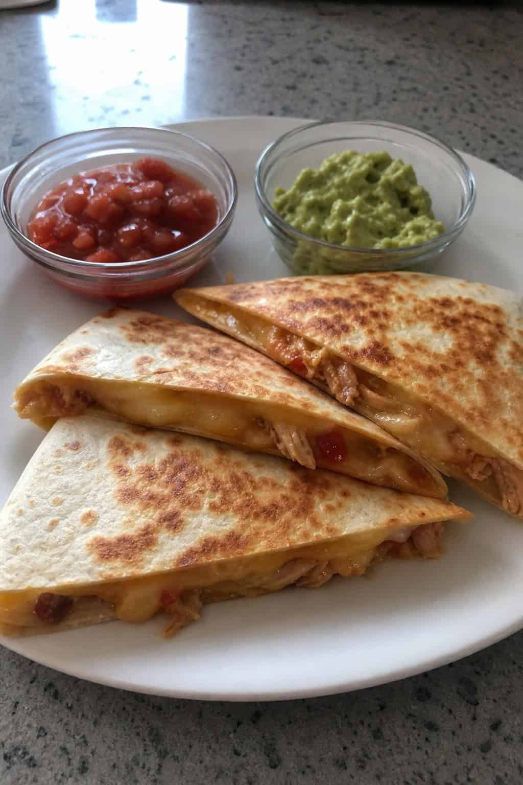 Three golden-brown cheesy chicken quesadilla triangles on a wooden board with bowls of salsa and guacamole.