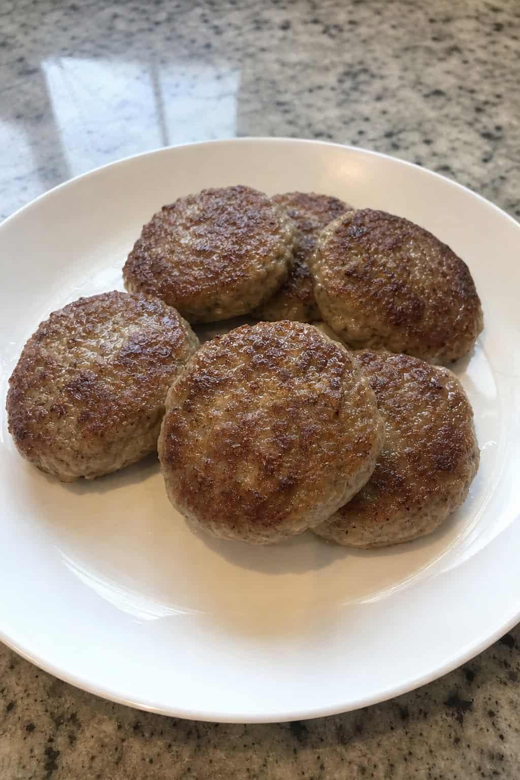 A plate of chicken and apple sausage patties garnished with herbs on a wooden table with apples and mustard sauce nearby.