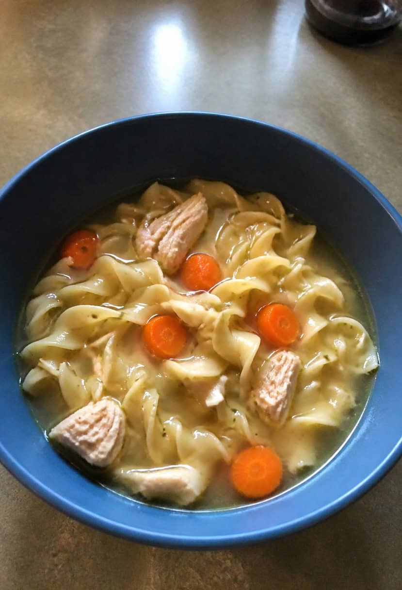 A steaming bowl of chicken noodle soup surrounded by fresh ingredients and a ladle