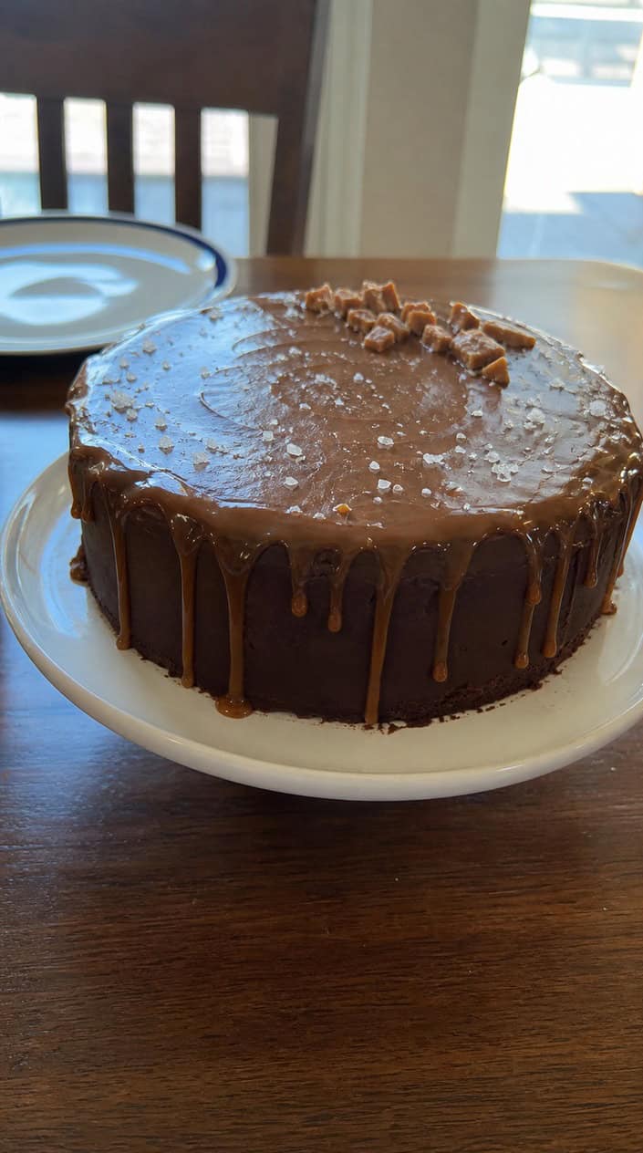 A chocolate ganache torte with sea salt caramel dripping down the sides, displayed on a white cake stand with sea salt flakes sprinkled around.