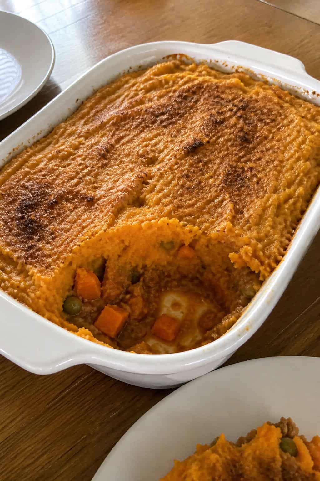 A freshly baked sweet potato shepherd's casserole in a ceramic dish on a wooden table with cinnamon sticks and sweet potatoes around it.
