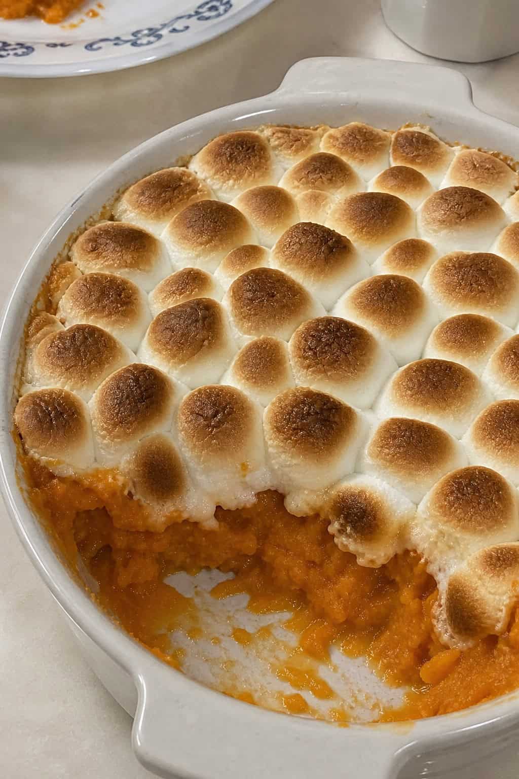 A sweet potato casserole topped with toasted marshmallows in a ceramic dish on a wooden table with autumn decorations around it.