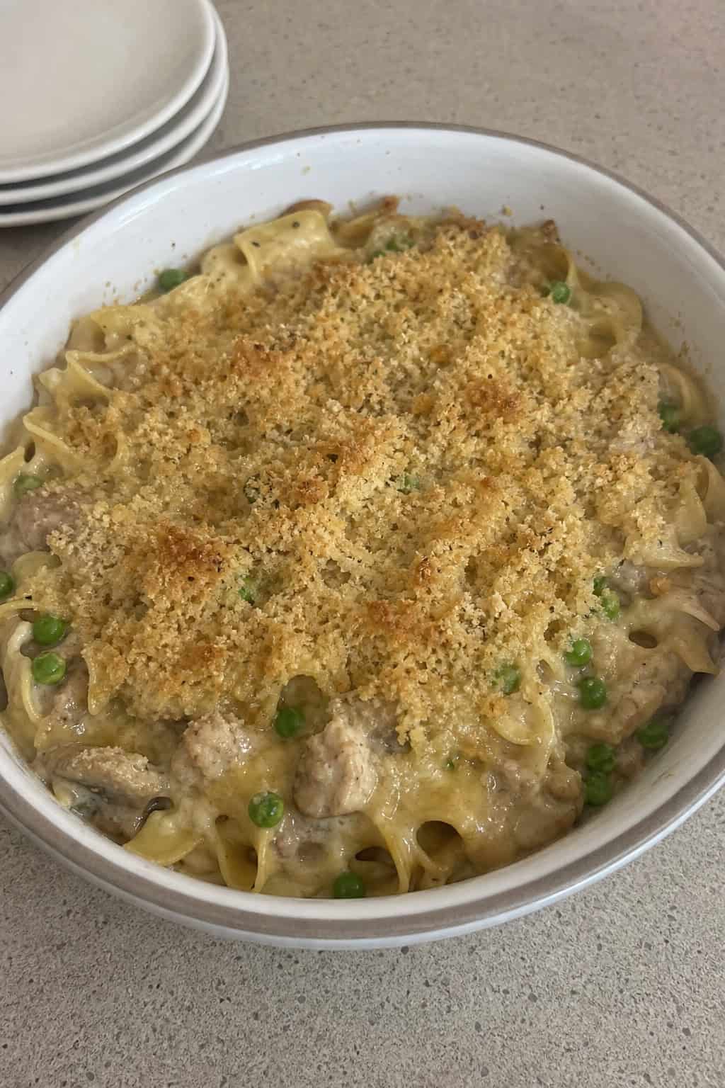 A baked tuna noodle casserole with egg noodles, cream sauce, and peas in a ceramic dish on a wooden table.