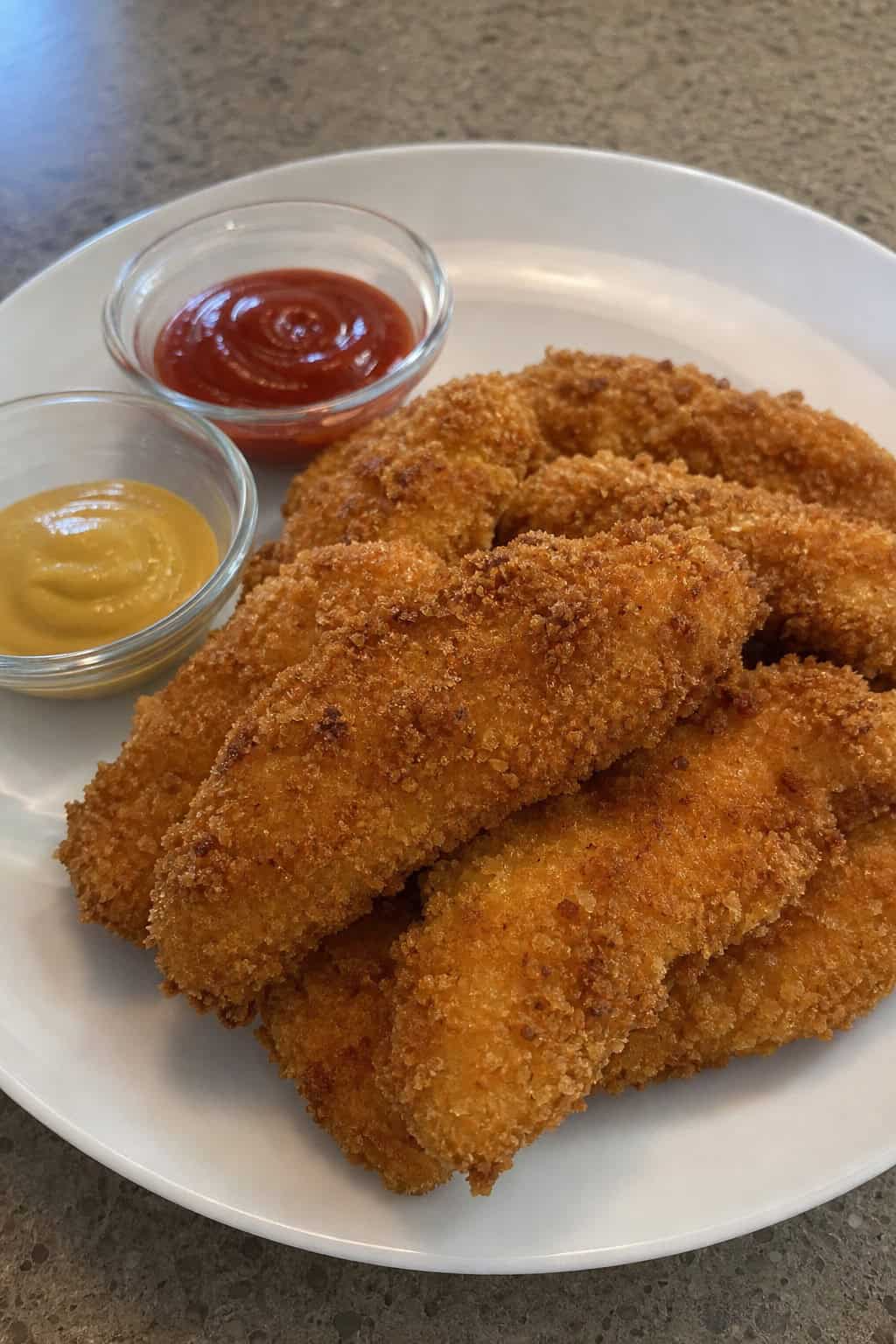 A plate of crispy oven-baked chicken tenders with dipping sauces on a wooden board.