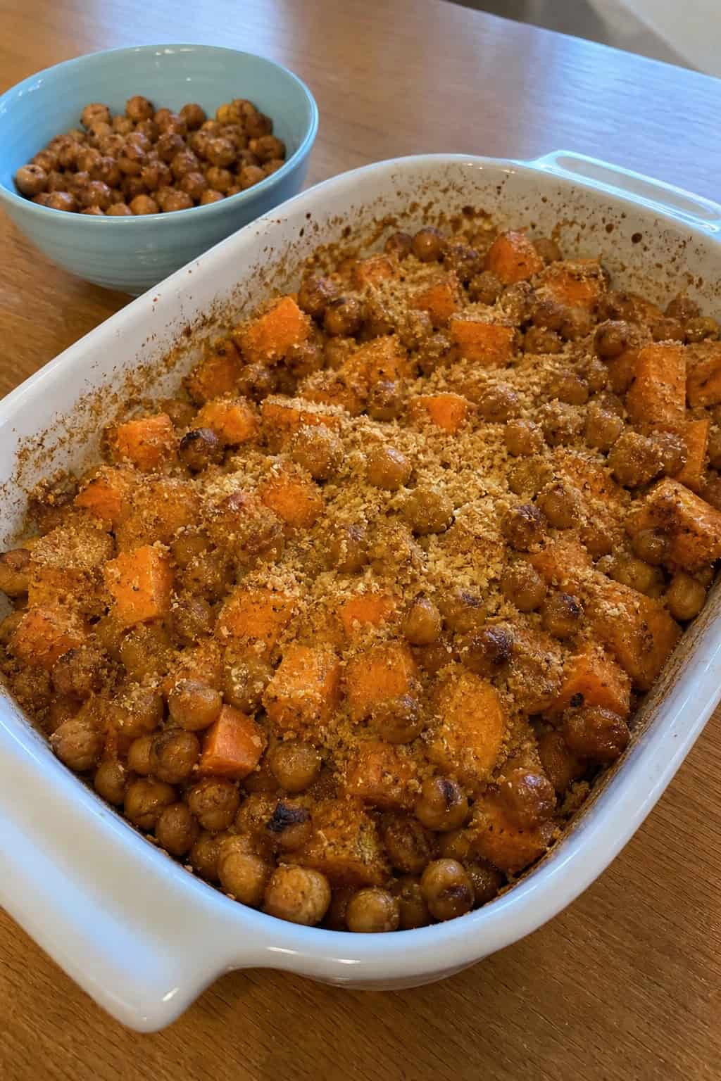 A baked sweet potato casserole topped with crispy chickpeas in a ceramic dish on a wooden table.