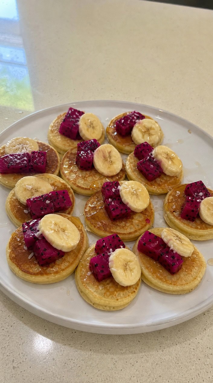 Small round pancake bites on a white plate showing flecks of bright pink dragonfruit and golden-brown edges, with a few tropical leaves tucked in the background.
