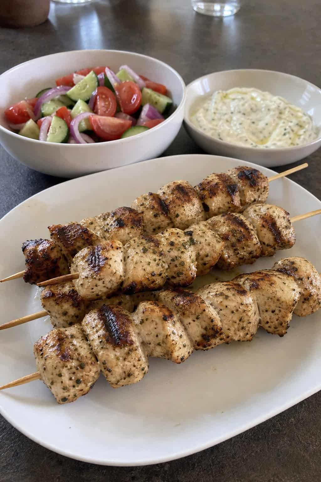 Grilled chicken skewers marinated in Greek yogurt served on a wooden board with fresh herbs and dipping sauces.