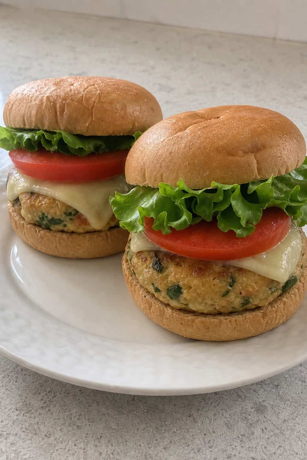Two chicken burgers with visible vegetables, lettuce, tomato, and cheese on a plate with sweet potato fries and dipping sauce on a kitchen table.