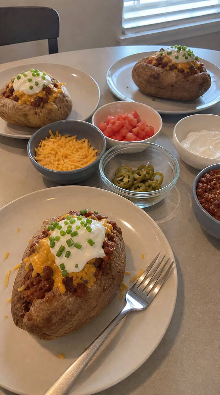 A table with baked potatoes topped with chili, cheese, sour cream, and chives, surrounded by bowls of various toppings for a game day meal.