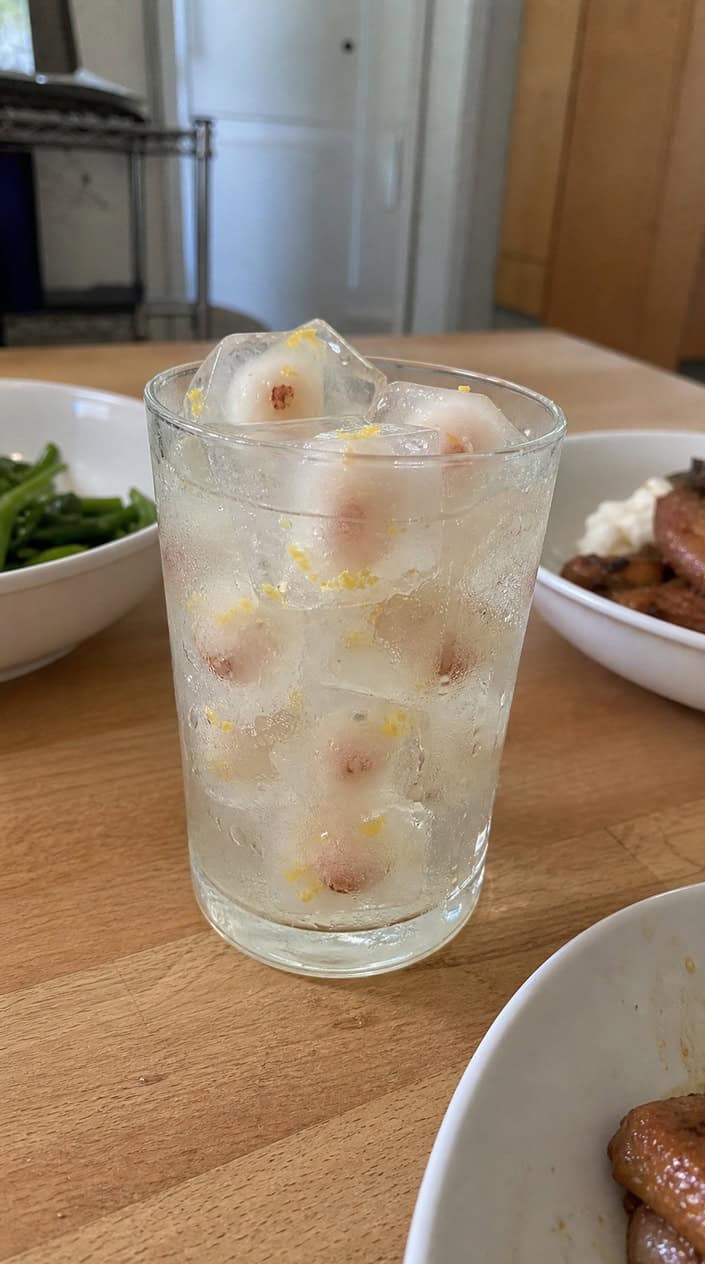 A clear glass with pale yellow lemonade and two lychee-studded ice cubes inside, fresh lychees and lemon wedges sitting on a slatted wooden surface beside it.