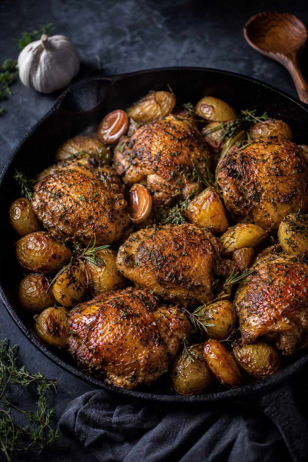 A well-seasoned cast-iron skillet holding four golden bone-in chicken thighs resting on halved baby potatoes, garnished with fresh thyme sprigs and minced parsley.