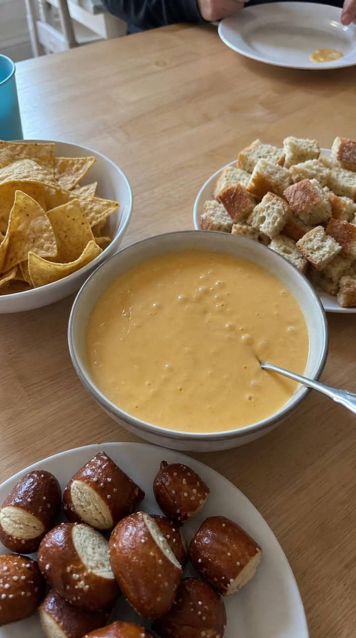 A bowl of creamy nacho cheese beer cheese fondue surrounded by tortilla chips, pretzel bites, vegetables, and bread on a wooden table.