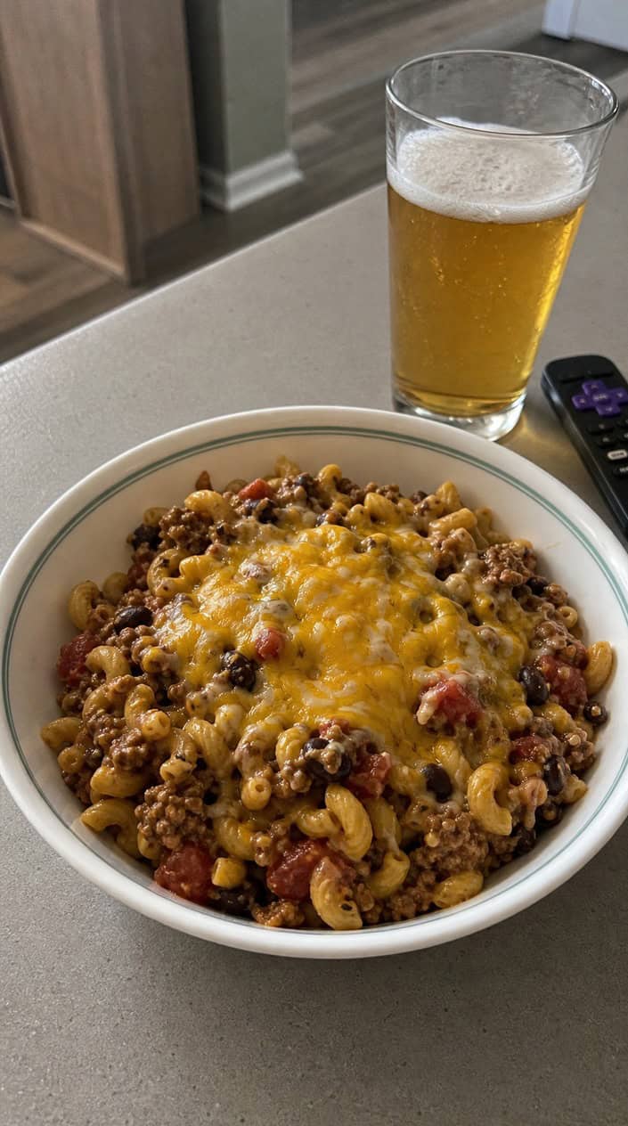 A bowl of cheesy taco pasta with ground beef, beans, corn, tomatoes, and fresh herbs on a wooden table with tortilla chips nearby.