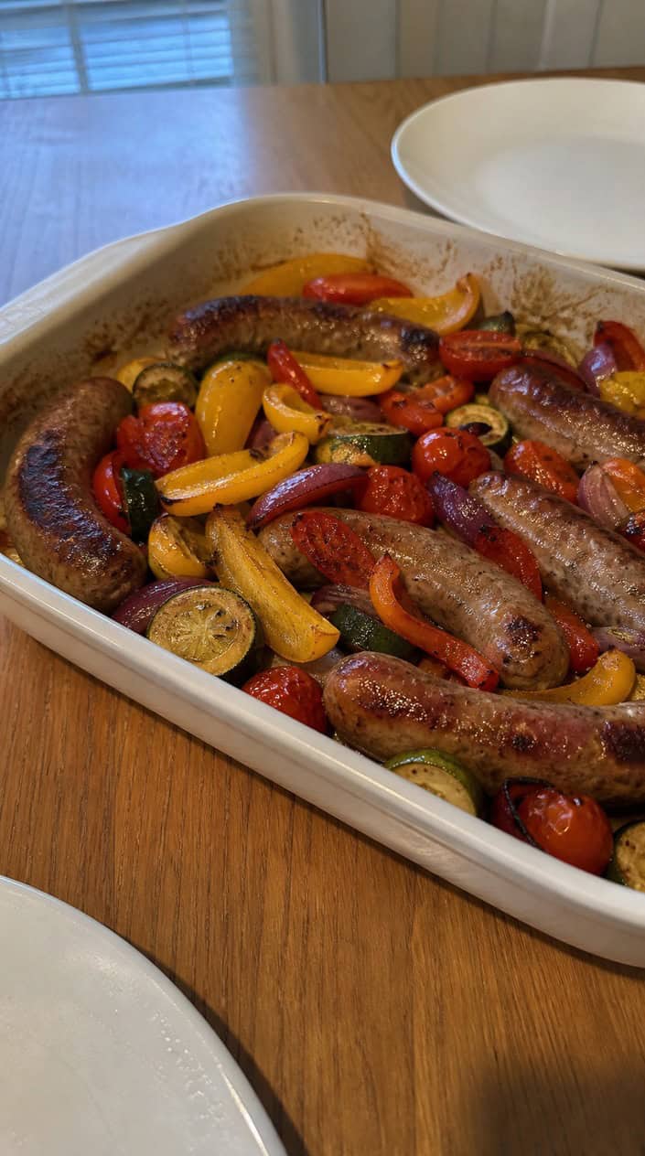 A plate of Italian sausage with roasted vegetables on a wooden table surrounded by small bowls of herbs and olive oil.
