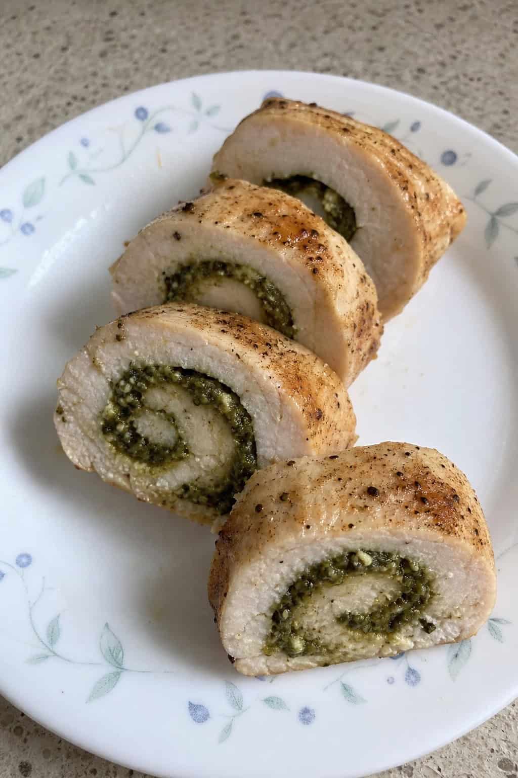 A platter of sliced pesto chicken roll-ups garnished with basil leaves and cherry tomatoes on a wooden table.