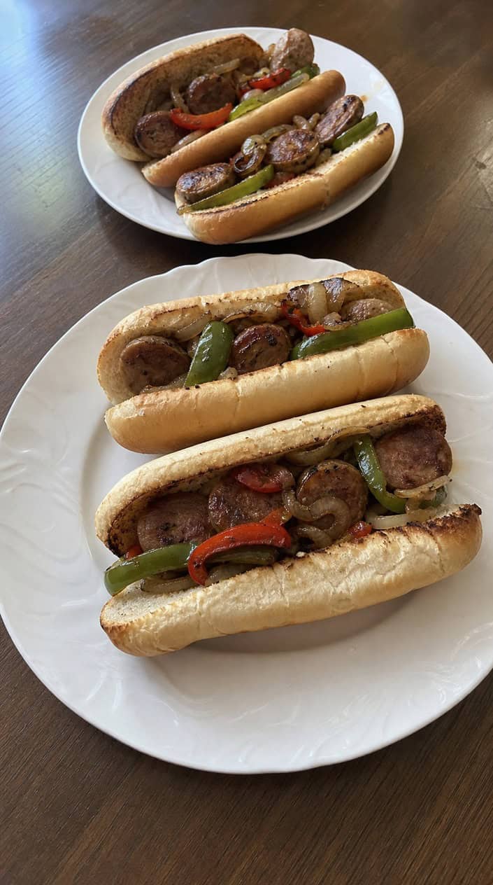 Several sausage, peppers, and onions hoagies on a wooden table with fresh ingredients in the background.