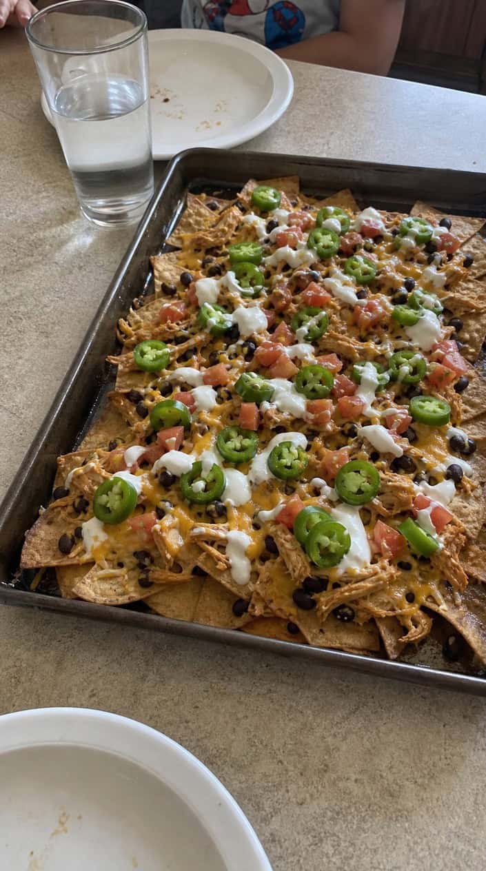 Sheet pan filled with loaded nachos topped with shredded rotisserie chicken and various toppings on a wooden table with bowls of salsa, guacamole, and sour cream nearby.